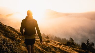 Eine Person wandert bei Sonnenaufgang auf einem grasbewachsenen Hügel mit Blick auf nebelbedeckte Wälder. | © Ferienregion Nationalpark Hohe Tauern - Gerald Demolsky