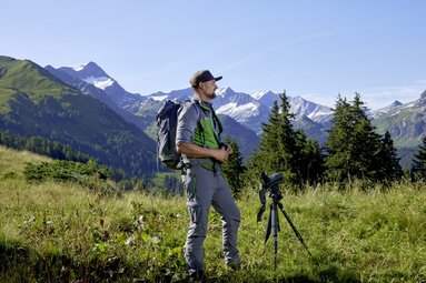 Ranger mit Fernglas und Stativ steht in einer Berglandschaft mit Wiesen und Blick auf schneebedeckte Gipfel. | © Ferienregion Nationalpark Hohe Tauern  - Michael Huber