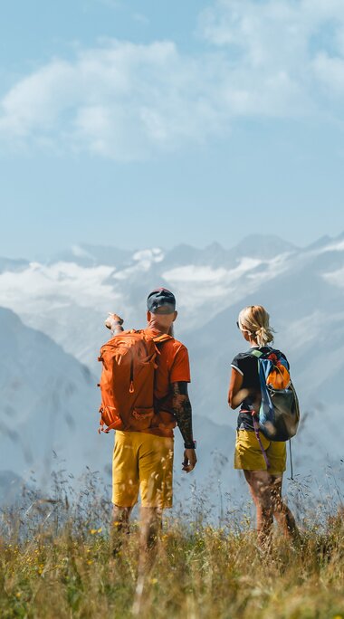 Zwei Wanderer auf einer Almwiese mit Blick auf schneebedeckte Berggipfel. | © Alpina Antica Trail